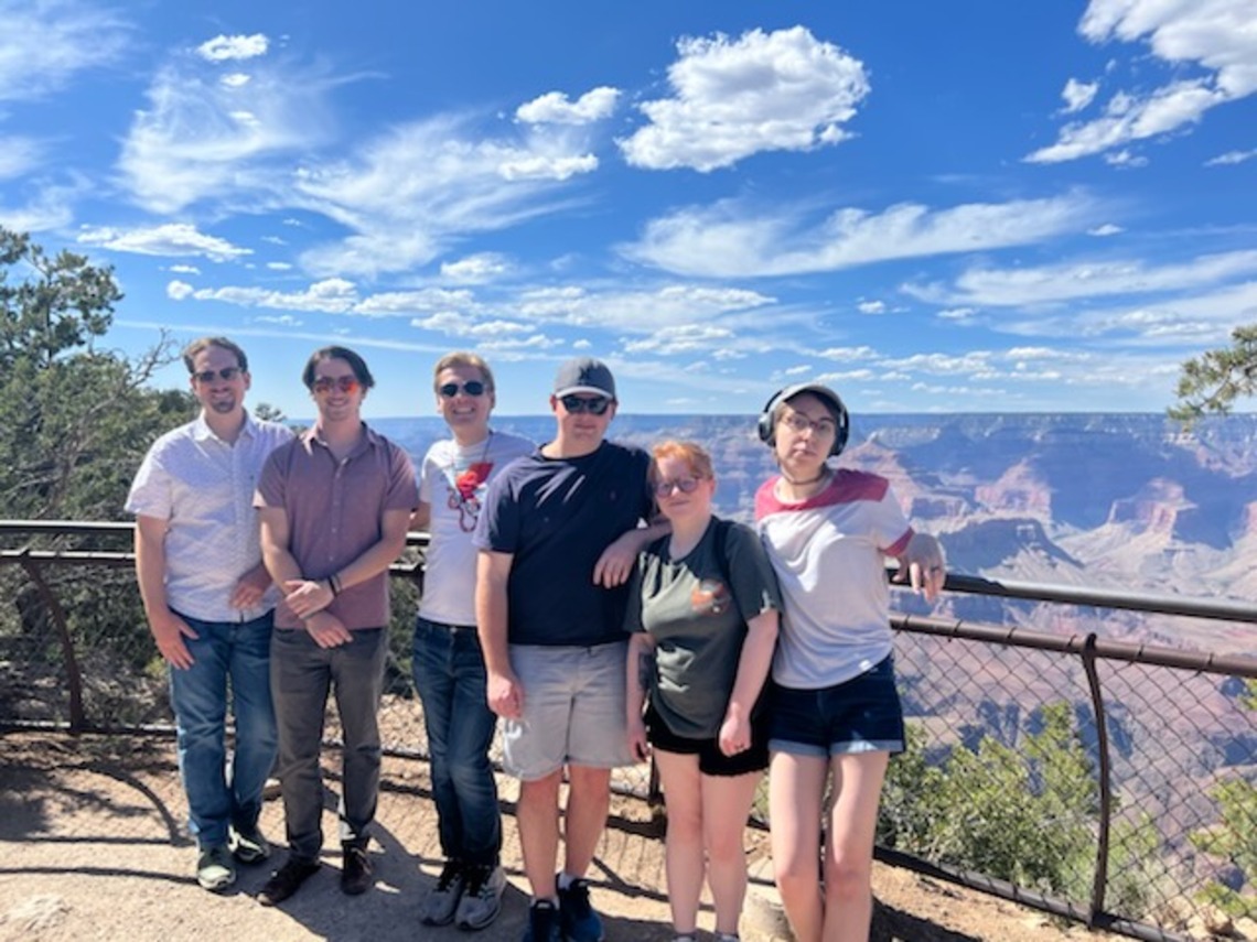 A group of 6 people standing in front of the grand canyon. The sky looks expansive and blue in the background.